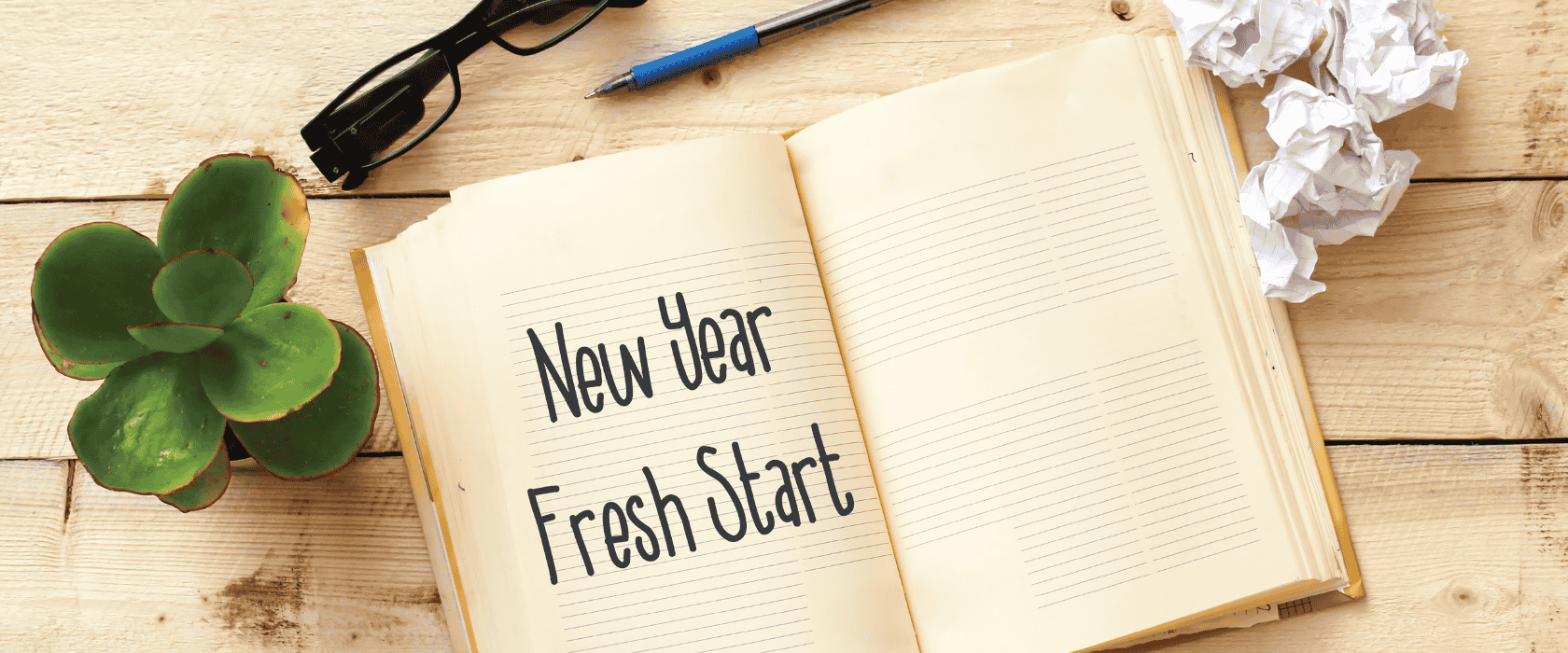 A journal with the words New Year Fresh Start written on it near a plant, glasses, a pen, and crumpled up paper.
