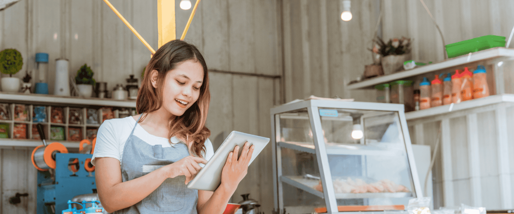 A young woman in a shop in an apron and holding a tablet in her hand.