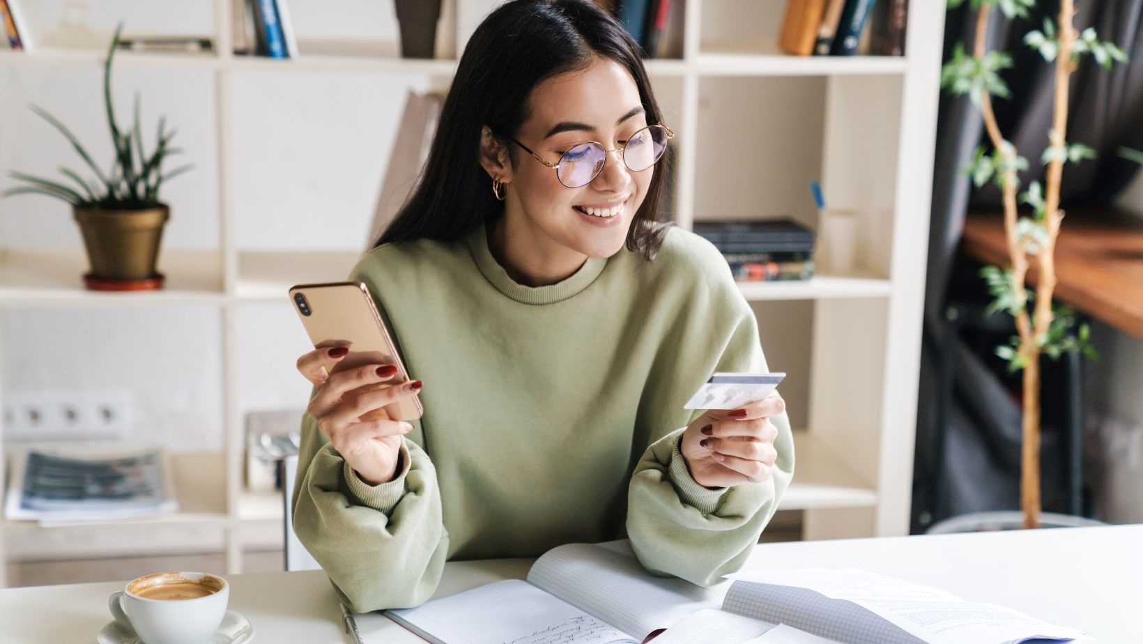 A young woman wearing glasses with her credit card in her hand and using her mobile phone to check her bank account.