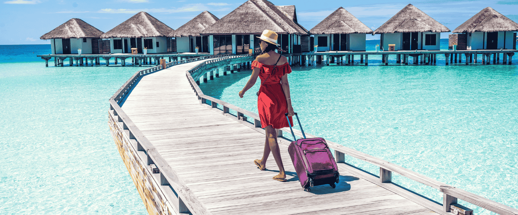 A woman walking on a raised boardwalk over water to huts over the water on vacation with a suitcase.